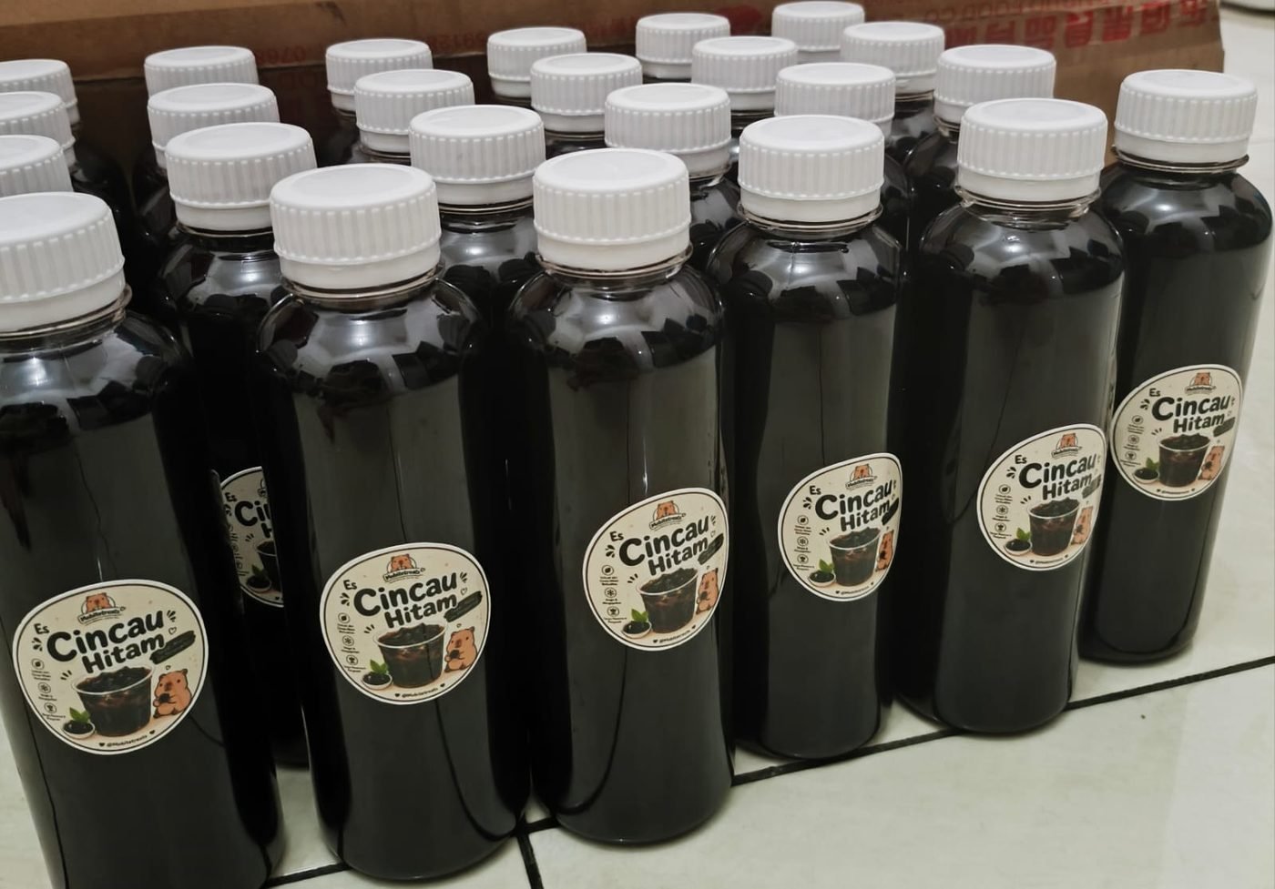 Deretan botol Es Cincau Hitam bertutup putih dengan stiker bulat bergambar kapibara