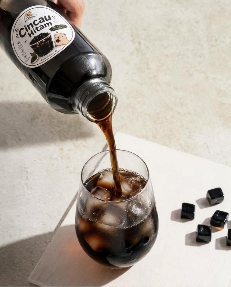 Es Cincau Hitam dituang dari botol ke gelas berisi es, potongan cincau hitam berserakan di meja